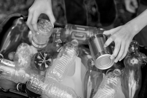 Close-up view of a person's hands sorting and collecting recyclable waste, including various plastic bottles and a metal can, which are being gathered together in a container or bin. The plastic bottles show a range of shapes and sizes, with some having visible caps and different textured surfaces, while the metal can has a smooth, reflective surface. The scene appears to take place outdoors, possibly in a garden or driveway area, indicated by the blurred background. The person's hands are actively engaged in handling the objects, with one hand holding a plastic bottle and the other gripping the metal can. This activity relates to rubbish collection and recycling, aligning with services provided by Garden Clearance Parsons Green, supporting environmentally responsible waste disposal in the Parsons Green area, SW6 postal code.