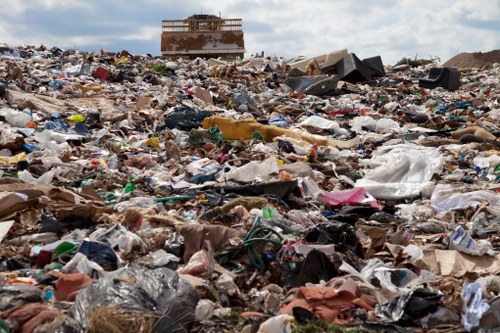 A vast expanse of open landfill site filled with a large accumulation of mixed waste materials, including plastic bags, cardboard, paper, textiles, and miscellaneous debris, spread across the ground in a densely packed, uneven layer. In the background, a partially constructed or abandoned building is visible under a cloudy sky, with the waste extending towards the horizon. Some waste items are visibly stuffed into black and white plastic bags, while other objects, such as broken wood, metal scraps, and discarded packaging, are scattered freely across the site. The scene depicts the environmental challenge of managing refuse in urban or semi-rural settings, which companies like Garden Clearance Parsons Green aim to address through professional rubbish removal services to help restore or maintain local areas.