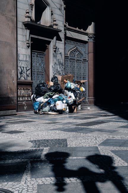 A large collection of mixed waste bags and discarded items piled against the exterior of a historic building with ornate, iron-barred windows and detailed stonework. The waste includes black rubbish b