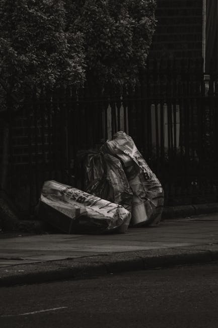 A large collection of mixed waste bags and discarded items piled against the exterior of a historic building with ornate, iron-barred windows and detailed stonework. The waste includes black rubbish b