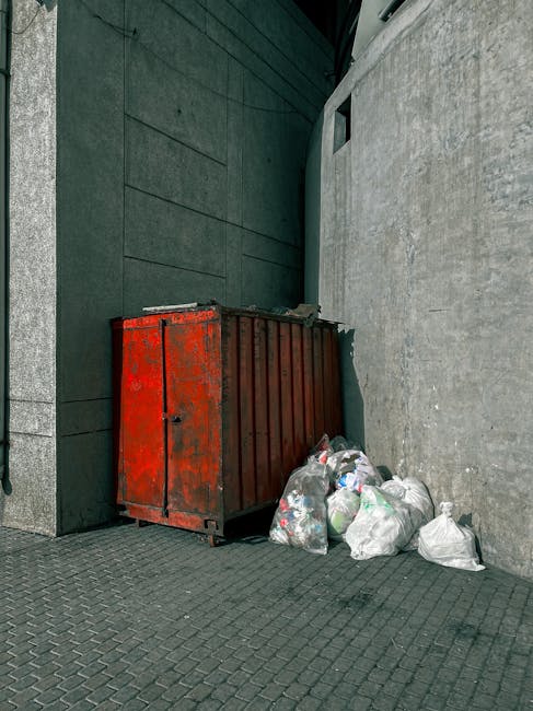 A sanitation worker in a yellow and red uniform is seen operating a large red rubbish collection vehicle on a street. The back of the vehicle has its hopper open, revealing mechanical components and i