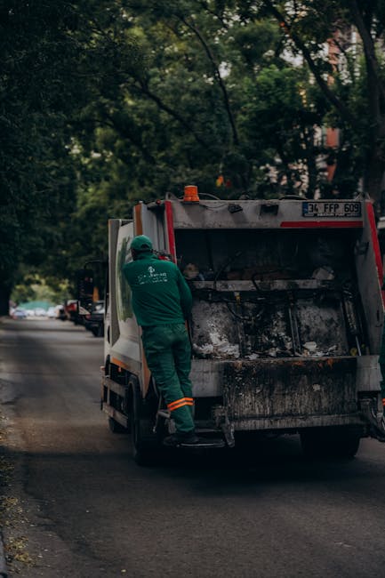 A small, old blue flatbed truck parked parallel to the street, heavily loaded with various types of waste materials for disposal. The truck's body shows signs of wear with faded paint and rusted red w
